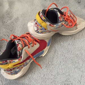 Nike Giannis Immortality färgglada sneakers - Nike Giannis Immortality sneakers med färgglatt bokstavsmönster, röd swoosh, gula och blå detaljer samt orange snören. Ovandel i syntet och textil, vit sula och svart innersida. Perfekta för dig som gillar unika och sportiga skor.