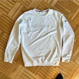 Vit långärmad Moncler tröja - Snygg vit långärmad tröja från Moncler med broderad logga på bröstet och patch på ärmen. Klassisk rund halsringning och mjukt bomullsmaterial. Perfekt för dig som gillar stilrena och exklusiva plagg med diskreta detaljer.
