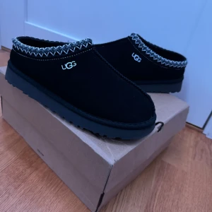 Svarta UGG slip-ons med mönstrad kant - Svarta slip-on skor från UGG med grov sula och snyggt mönstrad kant i grått och vitt. Skorna är i mocka och har UGG-logga på sidan. Perfekta för kalla dagar när du vill ha både stil och komfort. Skorna passar 42-44 eftersom de är lite för små 
