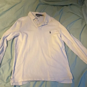 Vit långärmad pikétröja Polo Ralph Lauren - Vit långärmad pikétröja från Polo Ralph Lauren med klassisk krage och knäppning framtill. Tröjan har det ikoniska blå broderade logotypen på bröstet och är tillverkad i mjuk bomull. Perfekt för en clean och stilren look. Nypris är 1600kr