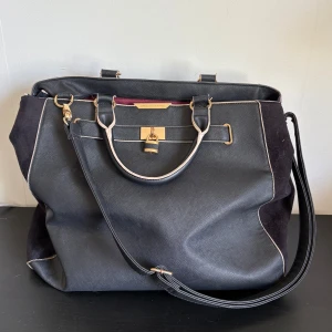 Stor svart handväska med gulddetaljer - Stilren svart axelväska med guldfärgade detaljer. Väskan har både handtag och lång axelrem, kontrasterande ljusa kanter och vinrött innerfoder. Väskan består av två stora fack med dragkedjor samt flera småfack! Perfekt till förvaring av dator eller anteckningsböcker! 