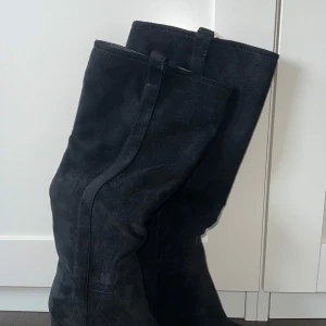 Svarta mockastövlar med klack - Snygga svarta stövlar i mocka med hög skaft och bred klack. De har rund tå och dragflikar upptill för enkel påtagning. Nyskick. Aldrig använda. Storlek 40.