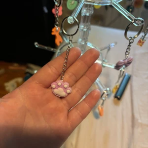 Sött nyckelring med tassberlock - Supergulligt nyckelring med en lila och vit tassformad berlock. Hängs på en tunn silverfärgad kedja. Perfekt accessoar för dig som älskar djur eller vill ha något lekfullt och unikt i din stil.