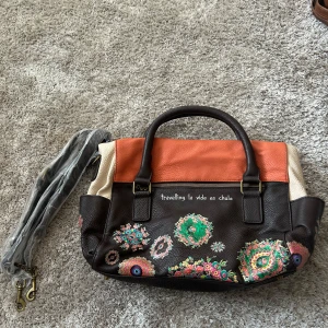 Desigual handväska med blommigt mönster - Säljer en unik handväska från Desigual i brunt, orange och beige skinn med färgglada blommiga detaljer. Väskan har både handtag och avtagbar axelrem, dragkedja och coolt mönstrat innerfoder. Texten 'travelling la vida es chula' broderad på framsidan.