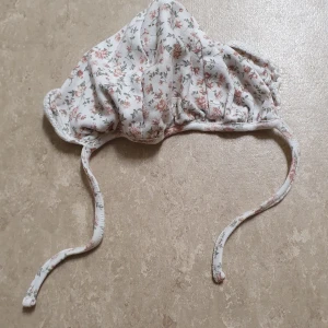 Mössa - Supersöt mössa från Newbie i mjuk bomull med blommigt mönster i rosa och grönt på vit botten. Mössan har knytband under hakan för extra bra passform och en klassisk form som täcker öronen.