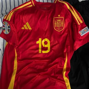Spanien Lamine Yamal matchtröja Adidas - Röd Spanien fotbollströja, nummer 19 och Lamine Yamal på ryggen. Klassisk Adidas-design med tre gula ränder på axlarna och landslagsemblem på bröstet. Tillverkad i lätt och ventilerande material, perfekt för fotbollsfans.