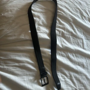 Svart flätat bälte med skinn-detaljer - Snyggt svart bälte med flätad design och detaljer i svart skinn vid spännet och änden. Spännet är fyrkantigt och matt svart, vilket ger ett stilrent intryck. Perfekt accessoar för att lyfta din outfit med en modern touch.