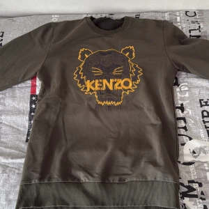 Olivgrön Kenzo sweatshirt med tiger - Säljer en olivgrön sweatshirt från Kenzo med ikoniskt broderat tigerhuvud och logga i gult och svart på bröstet. Tröjan har rund hals, ribbade muddar och är långärmad. Perfekt för dig som gillar streetstyle och vill sticka ut med en fet detalj framtill.