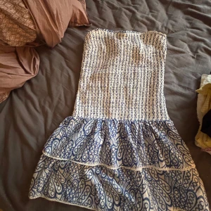 Vit och blå axelbandslös kortklänning - Supersöt axelbandslös kortklänning i vitt och blått med volanger och mönster. Smockad överdel och två lager volang i kjolen ger en luftig känsla.