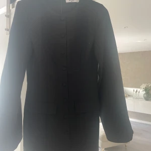 Svart klänning från NA-KD med knappar - Stilren svart klänning från NA-KD med långa ärmar och knappar framtill. Klänningen har en rak passform och två dekorativa fickor framtill. Perfekt för dig som gillar minimalistisk och klassisk stil.