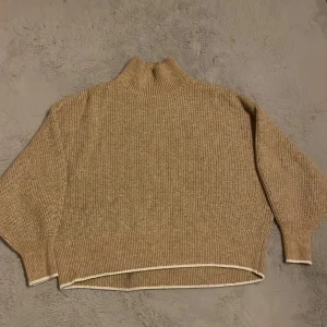 Beige stickad tröja - Tröjan har hög krage, lång ärm och en loose fit. Snygga vita detaljer längs nederkanten och ärmsluten. Den har en liten fläck på insidan (se bild) och en fläck som knappt syns på baksidan (den syns mer på bild, såg inte fläcken förens jag tog bilden)