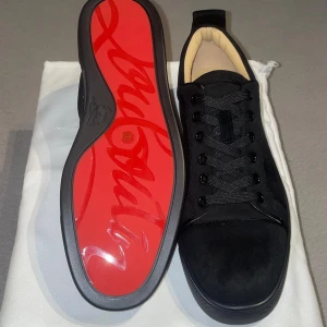 Svarta sneakers från Christian Louboutin - Snygga svarta sneakers från Christian Louboutin i mocka med klassisk röd sula och diskret logga på sidan. Skorna har svarta snören, rund tå och en stilren design som sticker ut med den ikoniska röda undersidan. Perfekta för dig som vill ha en lyxig och clean look.