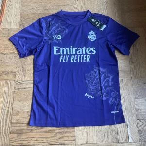 Lila Real Madrid x Y-3 fotbollströja - Snygg lila Real Madrid x Y-3 fotbollströja från Adidas med blommönster och tryckta loggor. Tröjan har korta ärmar, rund hals och är tillverkad i lätt, ventilerande material. Perfekt för dig som vill sticka ut på planen eller på läktaren.