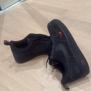 Nike Air Force 1 svart/röd mocka - Svarta Nike Air Force 1 sneakers i mocka med röda detaljer på logga, tunga och häl. Klassisk låg modell med snörning och rund tå. Snygg kontrast mellan svart och rött, perfekt för dig som gillar streetstyle och vill sticka ut, dessa är använda ett par gånger och slutade komma till använding då jag skaffa ett par andra, pris kan diskuteras ✅OBS!!! Dessa skor kan även passa 42-43