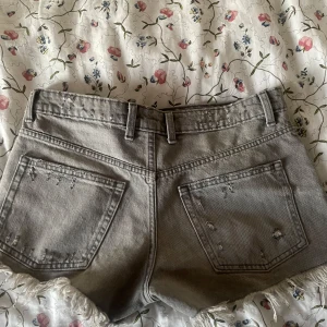 Grå jeansshorts med slitningar från Zara - Snygga grå jeansshorts från Zara med slitna detaljer och fransig kant nertill. Klassisk femficksmodell med knappar i gylfen och bälteshällor. Perfekta för en chill sommarlook med lite edge.