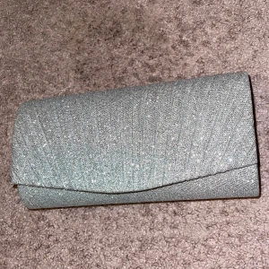 Glittrig silver kuvertväska - Snygg kuvertväska i glittrigt silver med elegant veckad design på framsidan. Perfekt accessoar för att lyfta din outfit med lite extra bling. Väskan har en stilren och modern form som passar till många olika looks.