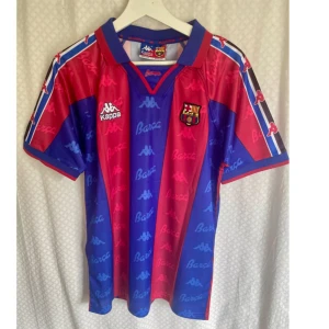 Vintage Jersey FC Barcelona - Klassisk fc Barcelona fotboll hemmatröja säsong 1993/1994 från Kappa med Romario och nummer 10 på ryggen. Klassiska blå och röda vertikala ränder, klubbmärke på bröstet och Kappa-loggor samt 'Barça'-text tryckt över hela tröjan. Kortärmad modell i glansigt, lätt polyestermaterial.