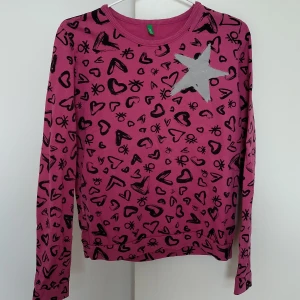 Rosa sweatshirt från Benetton - Rosa sweatshirt från United Colors of Benetton med svarta hjärtan och symboler över hela tröjan. Stor silverfärgad stjärna på bröstet. Långärmad och i mjuk bomull, perfekt för dig som gillar färg och mönster.