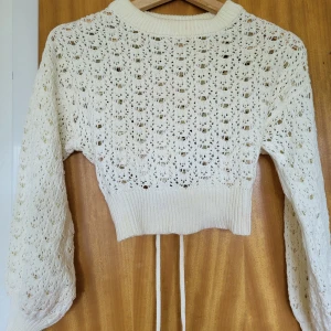 Vit stickad croppad kofta med snörning - Superfin croppad kofta i vit stickad bomull med hålmönster och långa ärmar. Koftan har snygg snörning i ryggen och bred ribbad mudd nedtill. 