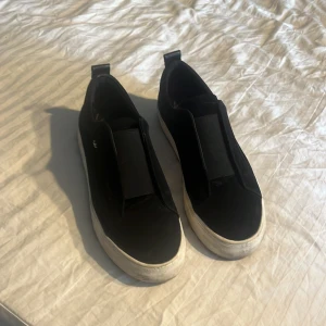 Svarta slip-on sneakers från KCobler - Knappt använda, storlek 37  svarta slip-on sneakers från KCobler med vit platt sula och elastiskt band över foten sulorna behöver bara tvättas. Skorna är i fau leather (tror jag) och har en minimalistisk look med diskret logga på sidan. Perfekta för dig som gillar simpel stil med snygga vardags skor. 