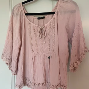 Rosa blus med spets från Elitzia - Supersöt ljusrosa blus från Elitzia med kort ärm och spetsdetaljer längs ärmslut och nederkant. Blusen har knytning framtill och en lös, flowy passform. Perfekt för dig som gillar romantisk och bohemisk stil. Notera den rosa fläcken på ärmen 