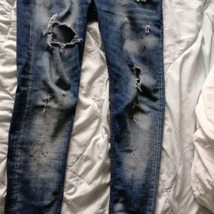 Dsquared2 slitna skinny jeans blå - Snygga blå jeans från Dsquared2 med skinny passform och slitna detaljer samt färgstänk för en edgy look. Klassisk femficksmodell med Dsquared2-logga på röd etikett och knapp. Perfekta för dig som gillar streetstyle och vill sticka ut.