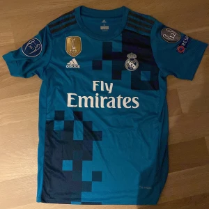Real Madrid Ronaldo bortatröja Adidas S - Säljer en blå Real Madrid bortatröja från Adidas med Ronaldo 7 på ryggen. Tröjan har korta ärmar, digitalt pixelmönster i mörkblått, FIFA och Champions League-märken på ärmarna samt Fly Emirates-tryck på bröstet. Tillverkad i lätt och ventilerande ClimaCool-material.