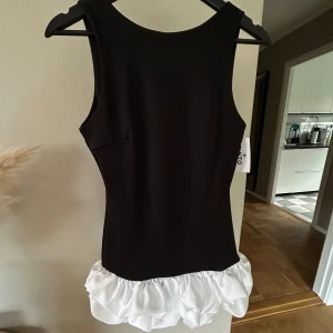 Svart miniklänning med vit volang och öppen rygg  - Otrolig miniklänning till festligare tillfällen! Sitter så snyggt på med perfekt längd. Helt ny, har prislappen kvar och är aldrig använd. Köpt i Spanien så går ej att få tag på här!