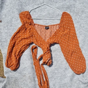 Prickig omlottopp från Gina Tricot - Superfin brun omlottopp från Gina Tricot med vita prickar. Toppen har långa puffärmar, v-ringning och knyt framtill. Perfekt för dig som gillar trendiga detaljer och en feminin look. Lätt och luftig i materialet, passar perfekt till jeans eller kjol.