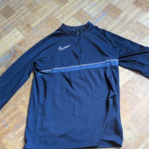 Nike Dri-Fit svart långärmad tröja XL - Svart långärmad Nike Dri-Fit tröja med halv dragkedja och vit broderad swoosh på bröstet. Tröjan har en tunn vit linje över bröstet och är tillverkad i ett lätt, syntetiskt material som andas. Perfekt för träning eller chill dagar. Det står XL men det känns som S.