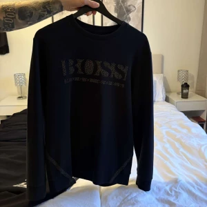 Svart sweatshirt från Hugo Boss XL - Svart sweatshirt från Hugo Boss i storlek XL. Tröjan har ett stort BOSS-tryck framtill i diskret mönster och rund halsringning. Långa ärmar och mjukt material som känns skönt mot huden. Perfekt för en clean och stilren look.