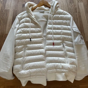 Moncler cardigan  - Säljer en snygg vit pufferjacka från Moncler med quiltad framsida, huva och dragkedja. Jackan har långa ärmar i mjuk bomull, två fickor med dragkedja och en Moncler-logga på ärmen. Perfekt för dig som vill ha en clean och stilren look.