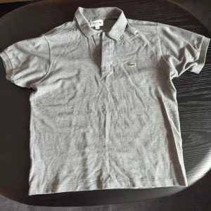 Grå pikétröja från Lacoste - Klassisk grå pikétröja från Lacoste med korta ärmar och den ikoniska gröna krokodilloggan på bröstet. Tröjan har krage och knäppning framtill. Perfekt för en clean och sportig stil.