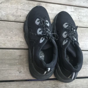 Svarta chunky sneakers med reflexdetaljer - Svarta chunky sneakers med grå och reflexdetaljer. Skorna har snörning med svarta och vita skosnören, grov sula och ovandel i textil och syntet. Perfekta för dig som gillar en sportig och trendig look.