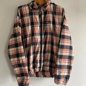 Rutig vindjacka från Patagonia - Snygg rutig vindjacka från Patagonia i blått, orange och vitt. Jackan har huva, hel dragkedja och elastiska muddar. Perfekt för dig som gillar streetstyle och vill ha något lätt och funktionellt. Klassisk logga på bröstet och coolt mönster som sticker ut.