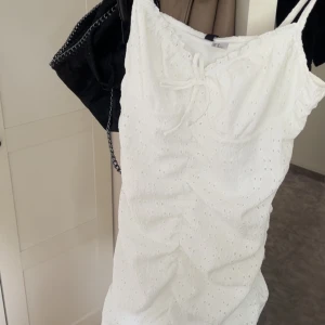 Vit sommarklänning från H&M - Supersöt vit kortklänning från H&M med broderade hål, smala axelband och knyt fram. Perfekt för varma dagar och har en feminin, lätt volangkant upptill. Luftig och skön i bomull, passar perfekt till sneakers eller sandaler. Helt ny med prislapp kvar. Strl M men passar mer S