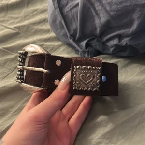 Brett brunt mockabälte med silverdetaljer - Säljer ett brett brunt bälte i mocka med ett hjärta på, detta är köpt på second hand och vet ej vilket märke det är. Den är ungefär 100cm lång 