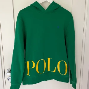 Grön hoodie från Polo Ralph Lauren - Snygg grön hoodie från Polo Ralph Lauren med stor gul POLO-text framtill. Klassisk huva, långa ärmar och fickor på sidorna. Tillverkad i mjuk bomull, perfekt för en chill och sportig look. Denna hoodie är i fint skick och är använd sparsamt. Säljer tröjan då den inte kommer till användning. 