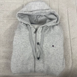 Grå hoodie från Tommy Hilfiger - Snygg ljusgrå hoodie från Tommy Hilfiger med dragkedja, huva med snörning och klassisk logga på bröstet. Mjuk bomullsblandning och ribbade muddar. Perfekt för chill dagar eller när du vill ha en clean och enkel look.passar M