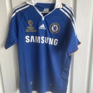 Chelsea Drogba 11 Champions League tröja - Chelsea FC fotbollströja från Champions League-finalen 2008 i Moskva. Blå med vita detaljer, Adidas-logga, klubbmärke och Samsung som sponsor. Nummer 11 och Drogba på ryggen. Klassisk krage med orange kant och vita ränder på axlarna. Tillverkad i lätt funktionsmaterial.