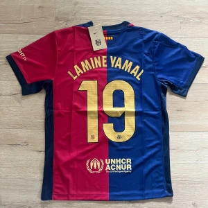 FC Barcelona Lamine Yamal Nike tröja - FC Barcelona matchtröja med Lamine Yamal 19-tryck. Halva tröjan är röd och halva blå, med guldiga detaljer och klubbmärke på bröstet. Snyggt Cactus Jack-tryck framtill och UNHCR-logga bak. Kortärmad modell i lätt, ventilerande material.