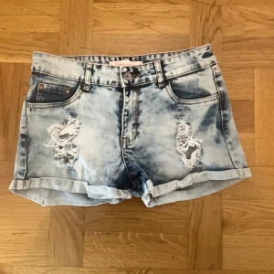 Blå slitna jeansshorts  - Snygga blå jeansshorts med slitna detaljer och uppvikta benslut. Shortsen har klassisk femficksmodell, bälteshällor och en cool tvättad look. Perfekta för sommardagar när du vill ha en avslappnad och trendig stil.