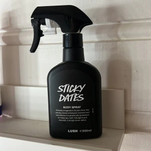 Sticky Dates Body Spray från Lush - Sticky Dates Body Spray från Lush i en svart plastflaska med spraymunstycke. Doften är söt och varm med inslag av benzoin och sandelträ. Flaskan rymmer 200 ml och har en modern, stilren design med vit text på svart bakgrund. Halva kvar 