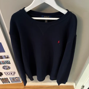 Mörkblå tröja från Polo Ralph Lauren - Klassisk mörkblå tröja från Polo Ralph Lauren med rund halsringning och röd broderad logga på bröstet. Tröjan har långa ärmar och är tillverkad i mjuk bomull, perfekt för en stilren och avslappnad look. Som ny!
