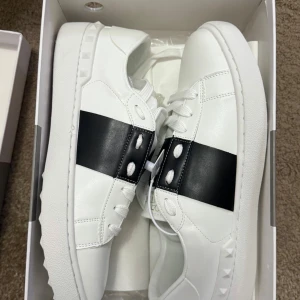 Valentino Garavani Open sneakers vit/svart - Snygga Valentino Garavani Open sneakers i vitt skinn med bred svart läderdetalj över mitten. Klassisk rund tå, vita snören och ikoniska nitar på hälen. Perfekta för dig som gillar stilrena och lyxiga sneakers med en twist.