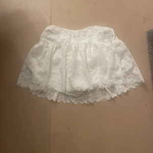 Vit spetskjol med volang - Supersöt vit kortkjol med spets och volangkant. Kjolen har inbyggda shorts under för extra komfort och säkerhet. Perfekt för dig som vill ha en romantisk och lekfull look. Spetsen ger en extra feminin touch och kjolen är lätt att matcha med olika toppar.