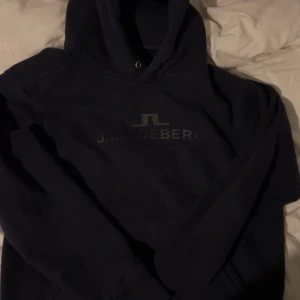 Mörkblå hoodie från J.Lindeberg - Säljer en stilren mörkblå hoodie från J.Lindeberg. Perfekt för en avslappnad look med sitt klassiska tryck på bröstet. Hoodien har en bekväm passform och är idealisk för kyligare dagar. Storlek L men är liten i storlek så skulle rekommendera M/S