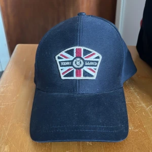 Mörkblå keps från Henri Lloyd - Snygg mörkblå keps från Henri Lloyd med broderad Union Jack-flagga och logga framtill. Klassisk böjd skärm och justerbar passform. Perfekt accessoar för en sportig och stilren look. Aldrig använd!!