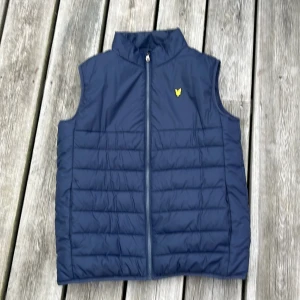 Marinblå dunväst från Lyle & Scott - Snygg marinblå dunväst från Lyle & Scott med gul logga på bröstet. Storlek 15/16 years och passar dig upp till 180 cm. Västen är använd ca 5 gånger. Säljer pga storlek. Bara att höra av sig vid frågor om västen💯Priset är inte hugget i sten.