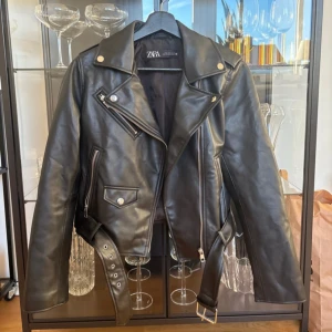 Svart skinnjacka från Zara med bälte - Snygg svart skinnjacka från Zara med klassisk bikerstil, silvriga dragkedjor, fickor och bälte i midjan. Jackan är i storlek M men passar mig som har XS